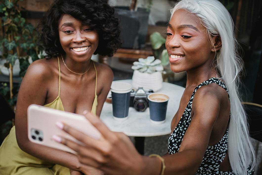 Two girls holding a phone, one with a long white blonde wig and the other with a short curly wig